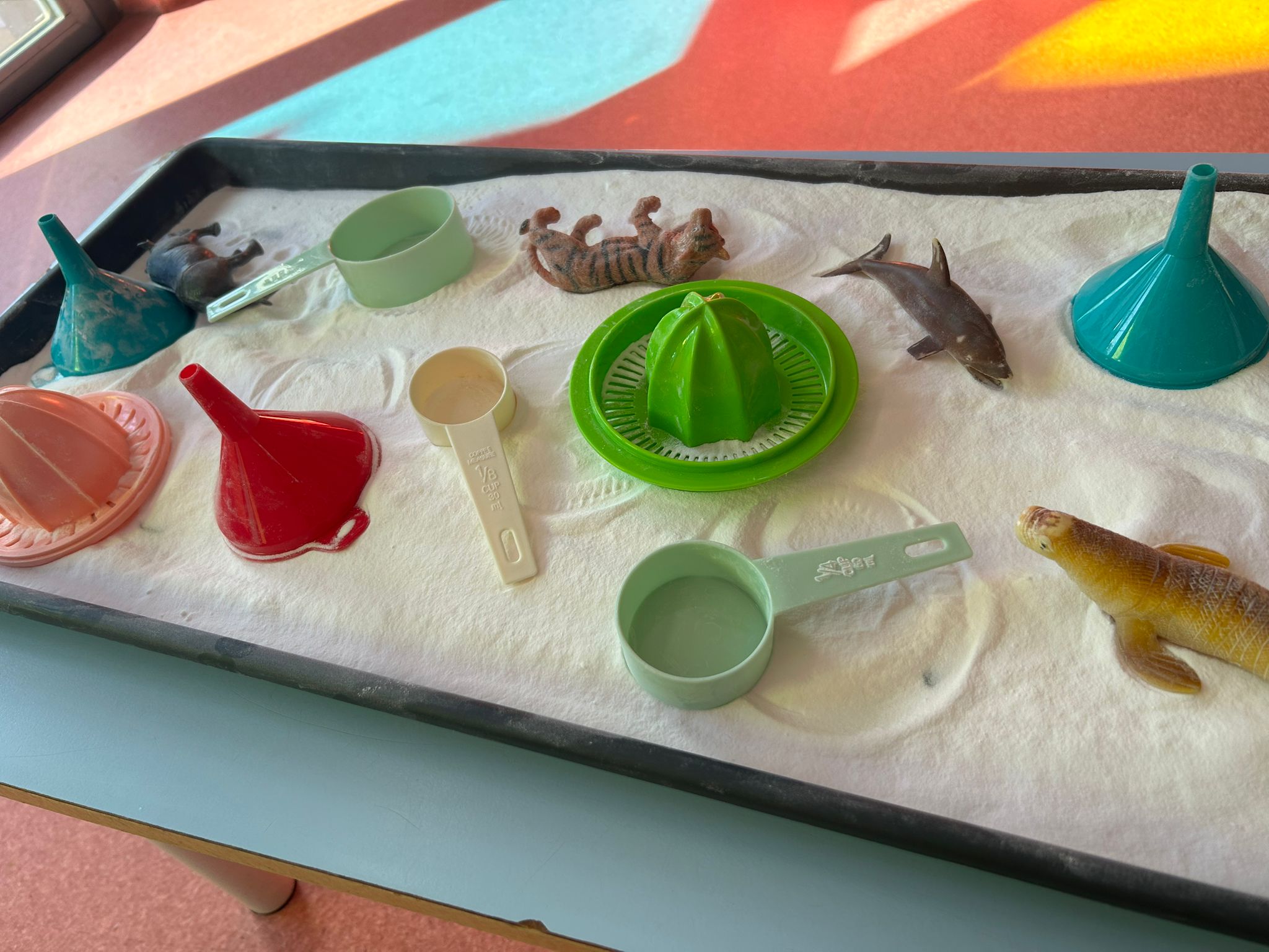 Bandeja preparada con harina, exprimidores de colores, embudos y animales de juguete para actividades infantiles.